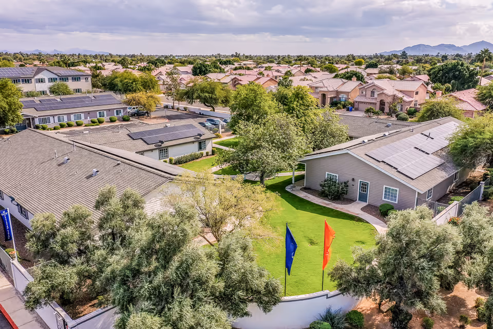 Aerial view of a senior living facility with multiple single-story buildings featuring solar panels on the roofs, surrounded by green lawns, trees, and a white perimeter wall. Residential houses and mountains are visible in the background under a partly cloudy sky.