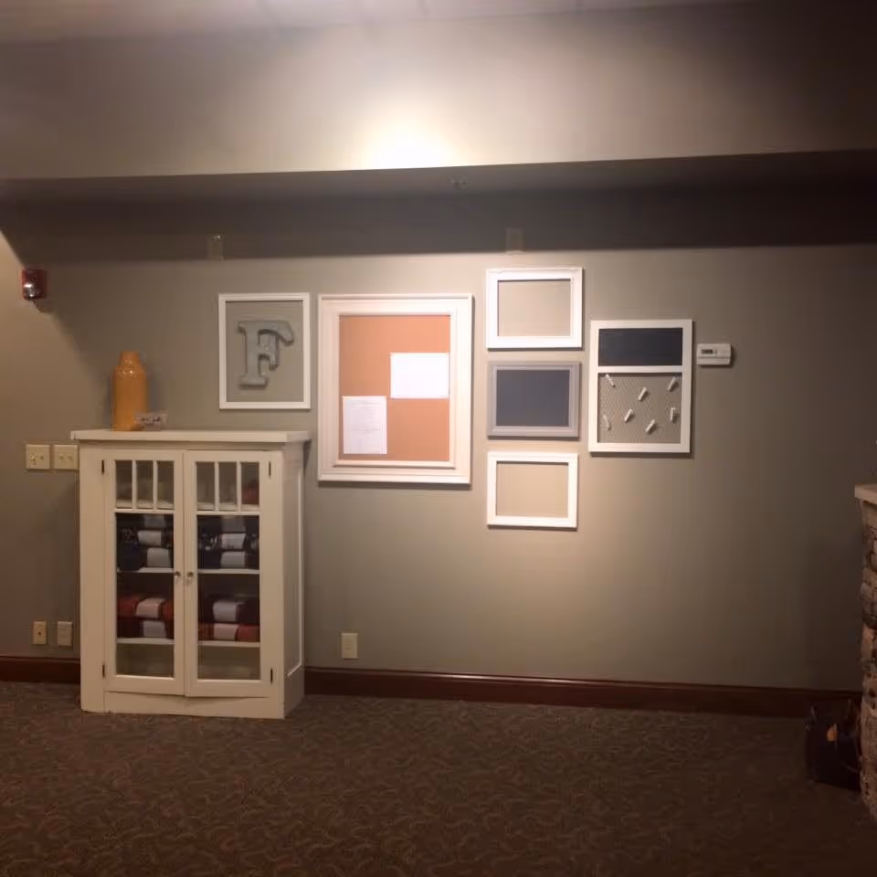 Interior wall of a senior living facility with a white cabinet containing folded blankets, a large yellow decorative vase on top, and several framed bulletin boards and decorative frames arranged on the wall above the cabinet.