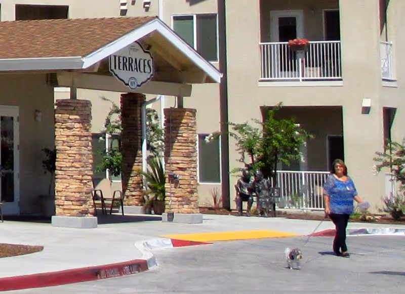 Entrance of a retirement community building with a covered portico labeled 'Terraces', stone columns, balconies, and a woman walking a small dog in front.