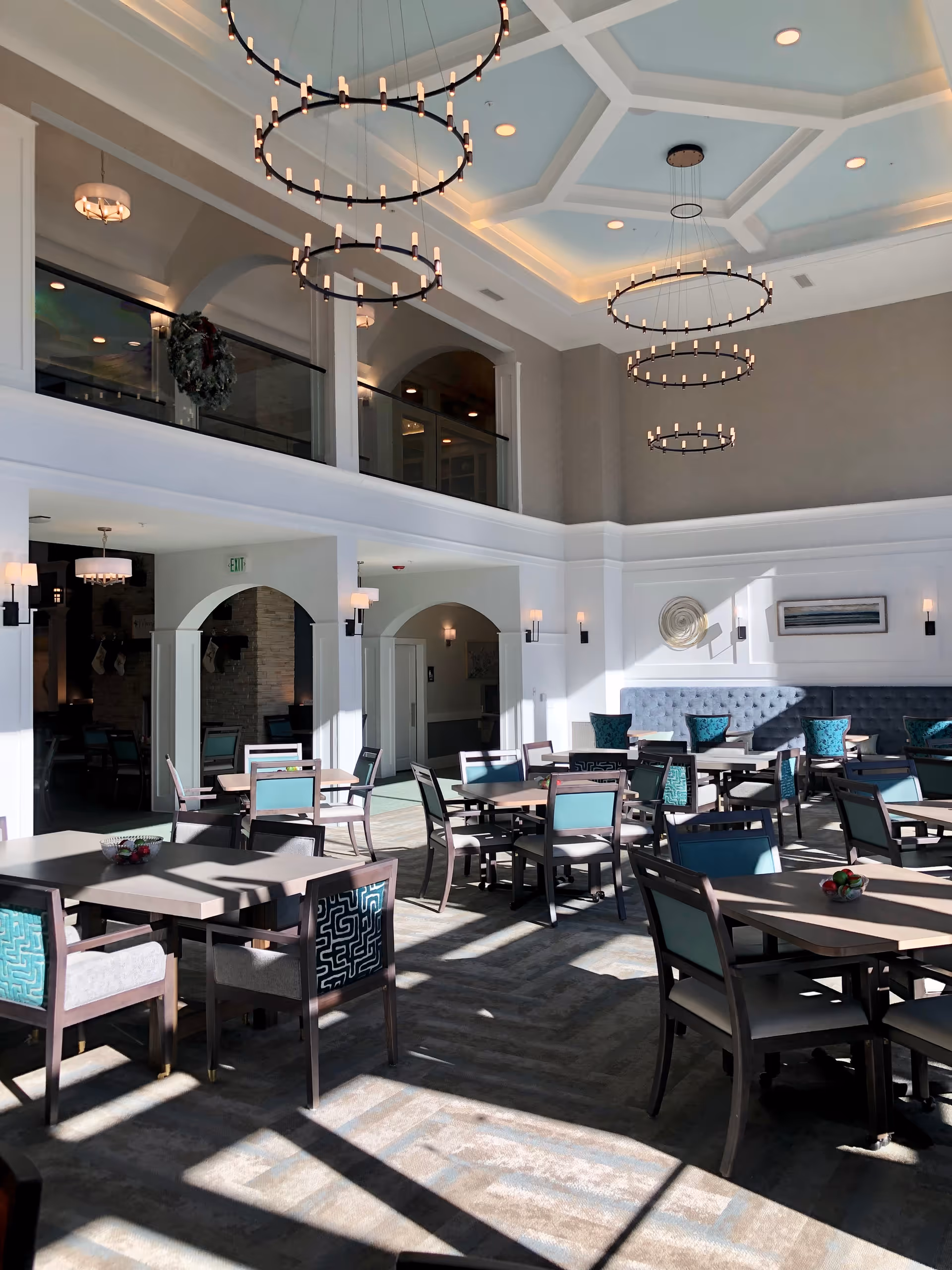 A bright and spacious dining room with multiple tables and chairs arranged neatly. The room features high ceilings with elegant circular chandeliers hanging down, large windows allowing natural light to fill the space, and a mix of teal and patterned upholstery on the chairs. There are wall sconces and decorative art pieces on the walls, with an upper balcony area visible.
