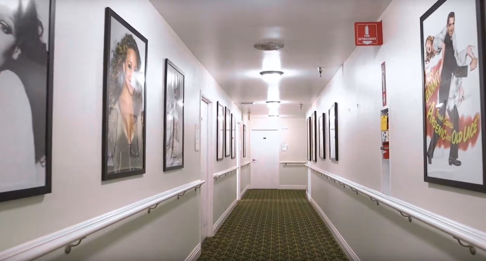A long, narrow hallway with green patterned carpet and white walls. The walls are decorated with framed black and white and color photographs or posters. Handrails run along both sides of the hallway. Ceiling lights illuminate the corridor, and a fire extinguisher is mounted on the right wall near the end of the hallway.