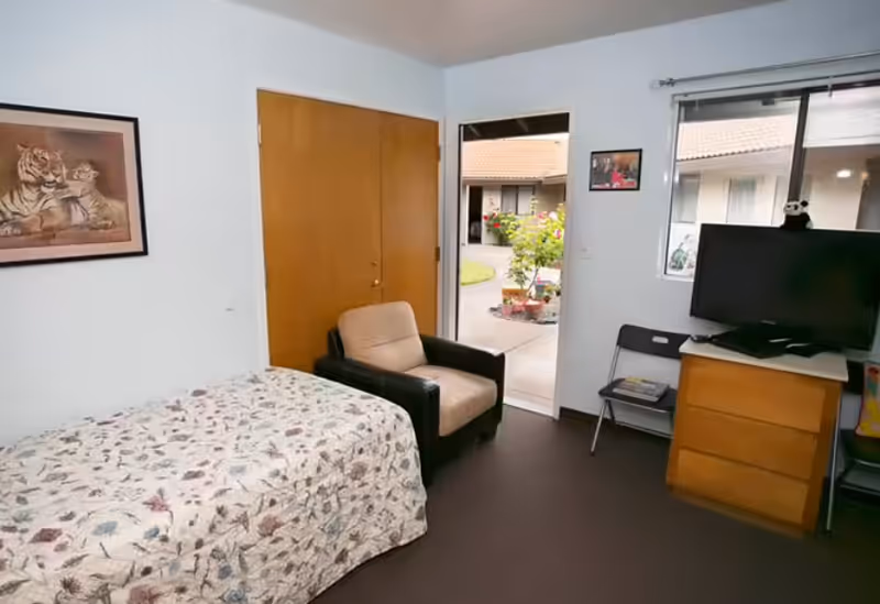 A simple bedroom with a single bed, armchair, dresser with a TV, and an open door and window looking onto a courtyard.