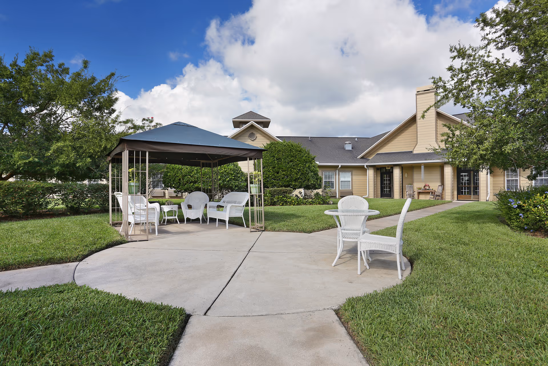 Outdoor seating area with a gazebo, white wicker chairs and tables on a paved patio in front of a beige senior living building.