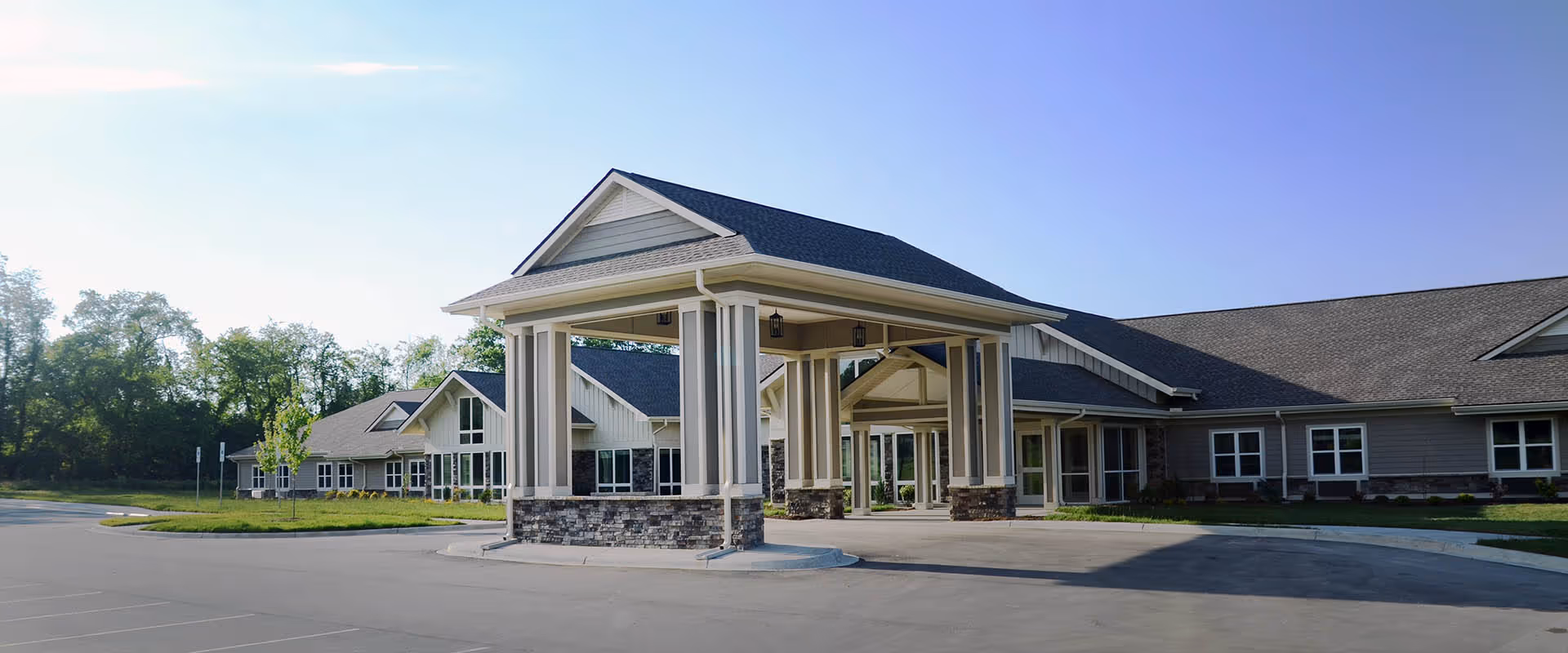 Exterior view of The Landings of Mills River senior living facility showing a large covered entrance with stone and siding accents, surrounded by a paved driveway and parking area, with trees and greenery in the background under a clear blue sky.