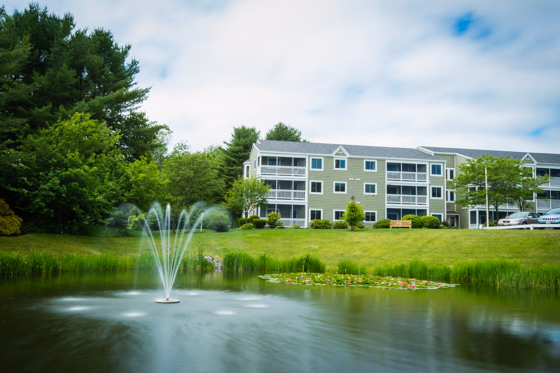 A serene outdoor scene at The Highlands featuring a pond with a water fountain spraying water upwards, surrounded by green grass and trees. In the background, there is a three-story residential building with balconies and parked cars nearby under a partly cloudy sky.