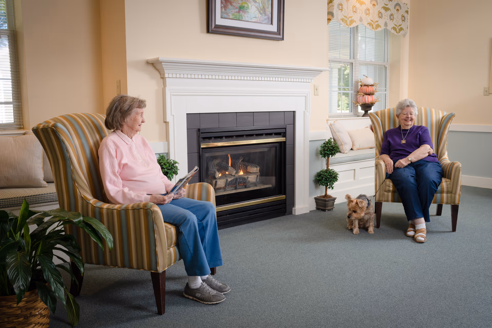Two elderly women sitting in striped armchairs facing each other in a cozy room with a lit fireplace between them. One woman is wearing a pink sweater and blue pants, holding a magazine, while the other woman is wearing a purple top and blue pants, smiling. A small dog is sitting on the carpet near the woman in purple. The room has light-colored walls, a window with floral curtains, a cushioned bench with pillows, potted plants, and a framed painting above the fireplace.