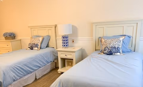 A bedroom with two twin beds, each with light blue bedding and decorative pillows. Between the beds is a white nightstand with a blue and white patterned lamp. There is a small dresser with a flower arrangement on the left side of the room. The walls are painted a light beige color.
