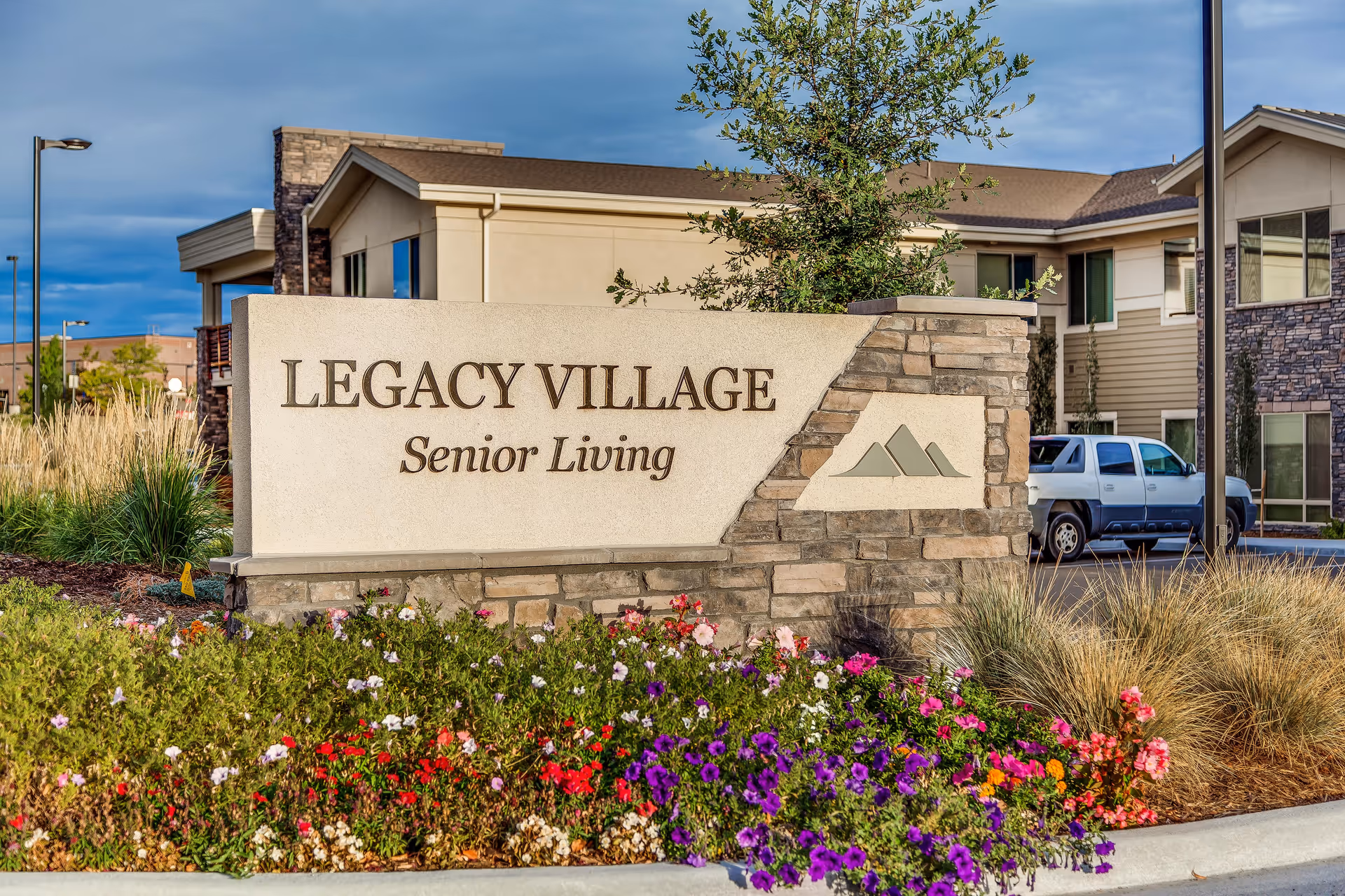 Stone and stucco sign for Legacy Village Senior Living surrounded by colorful flowers and landscaping, with part of the senior living facility building and a parked truck visible in the background under a partly cloudy sky.