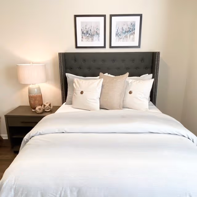 A neatly made bed with a dark upholstered headboard featuring button tufting and nailhead trim. The bed has white and beige pillows arranged against the headboard. On the left side, there is a wooden nightstand with a ceramic lamp and decorative objects. Two framed abstract art pieces hang on the wall above the bed.