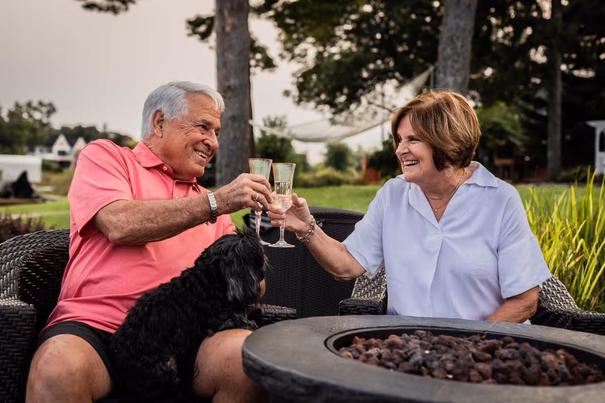 An elderly man and woman sitting outdoors on wicker chairs, clinking glasses of champagne and smiling at each other. A small black dog is sitting on the man's lap. They are surrounded by greenery and trees with a fire pit in the foreground.