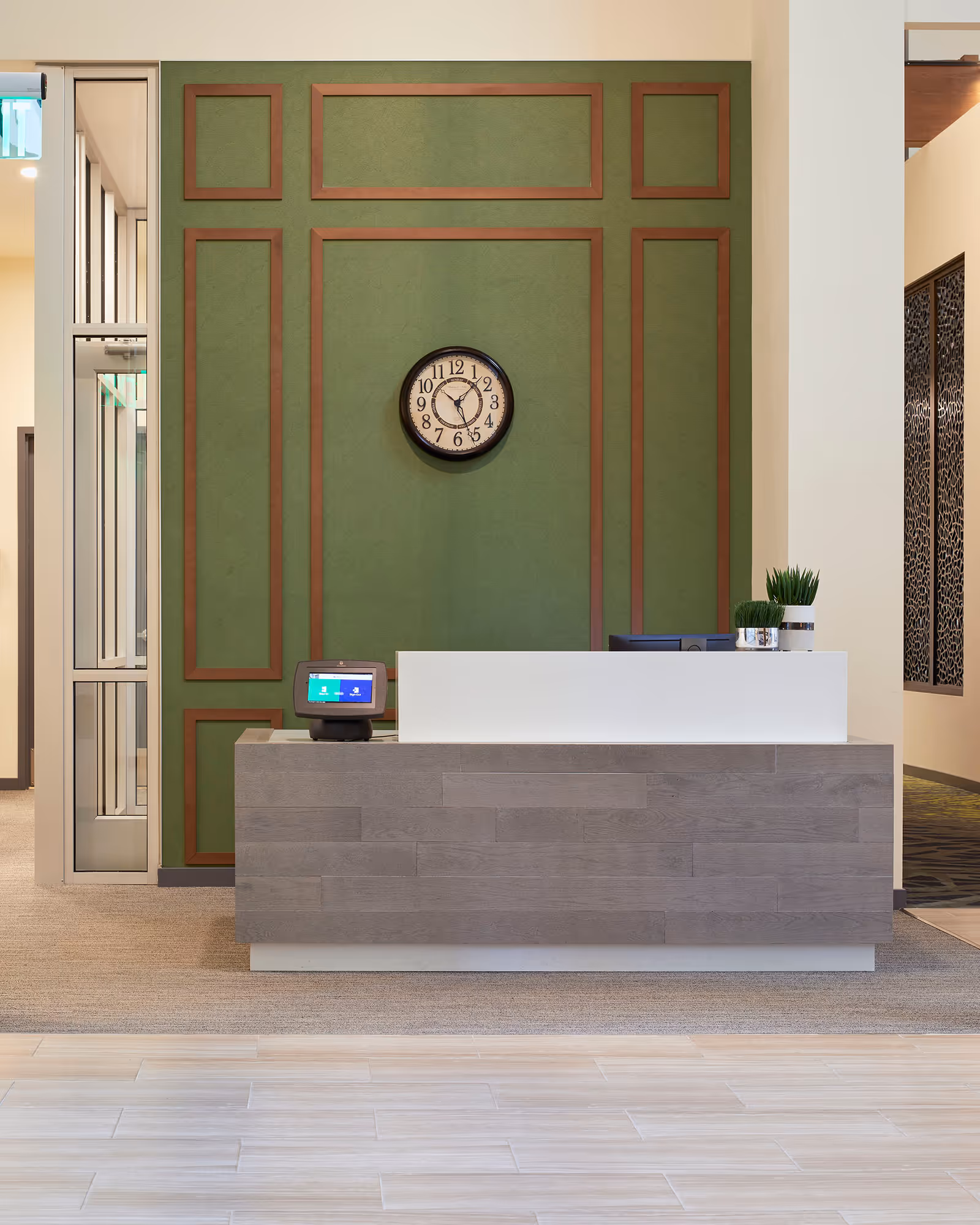 Reception desk area with a modern design featuring a wooden front panel and a white countertop. Behind the desk is a green wall with brown trim accents and a round clock showing the time as 6:30. There are two small potted plants on the desk and a digital device on the left side. The floor has light-colored tiles in the foreground and carpet behind the desk.