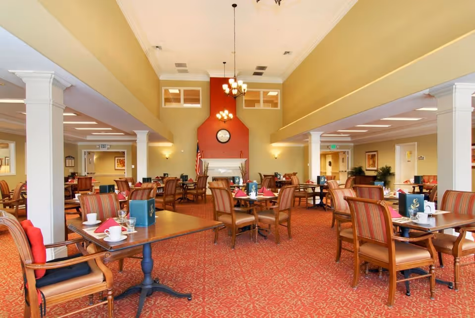 Spacious senior dining room with multiple tables and chairs, red patterned carpet, a fireplace wall with a clock and chandeliers.