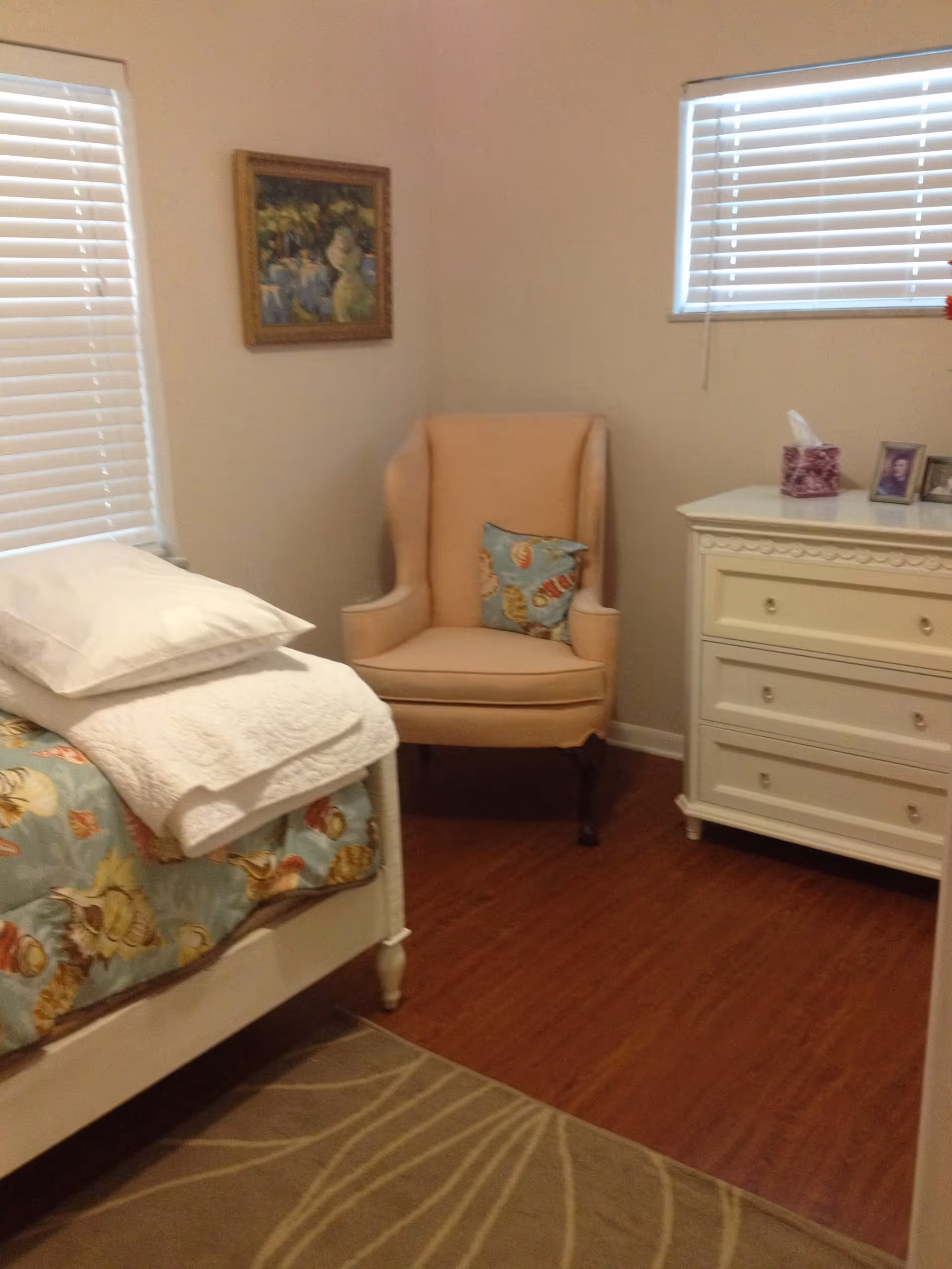 A small bedroom with a single bed covered with a seashell-patterned bedspread and white pillows and blanket. There is a beige upholstered armchair with a seashell-patterned cushion in the corner. A white dresser with decorative trim holds a tissue box and framed photos. Two windows with white blinds let in natural light. A framed painting hangs on the wall above the chair. The floor is wood with a beige area rug featuring a leaf pattern.