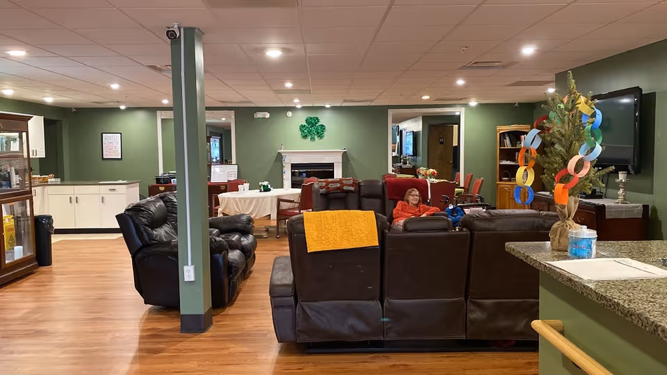 A communal living room with leather sofas arranged facing a fireplace, a seated resident, dining table, and a colorful paper-chain decoration on a small tree.