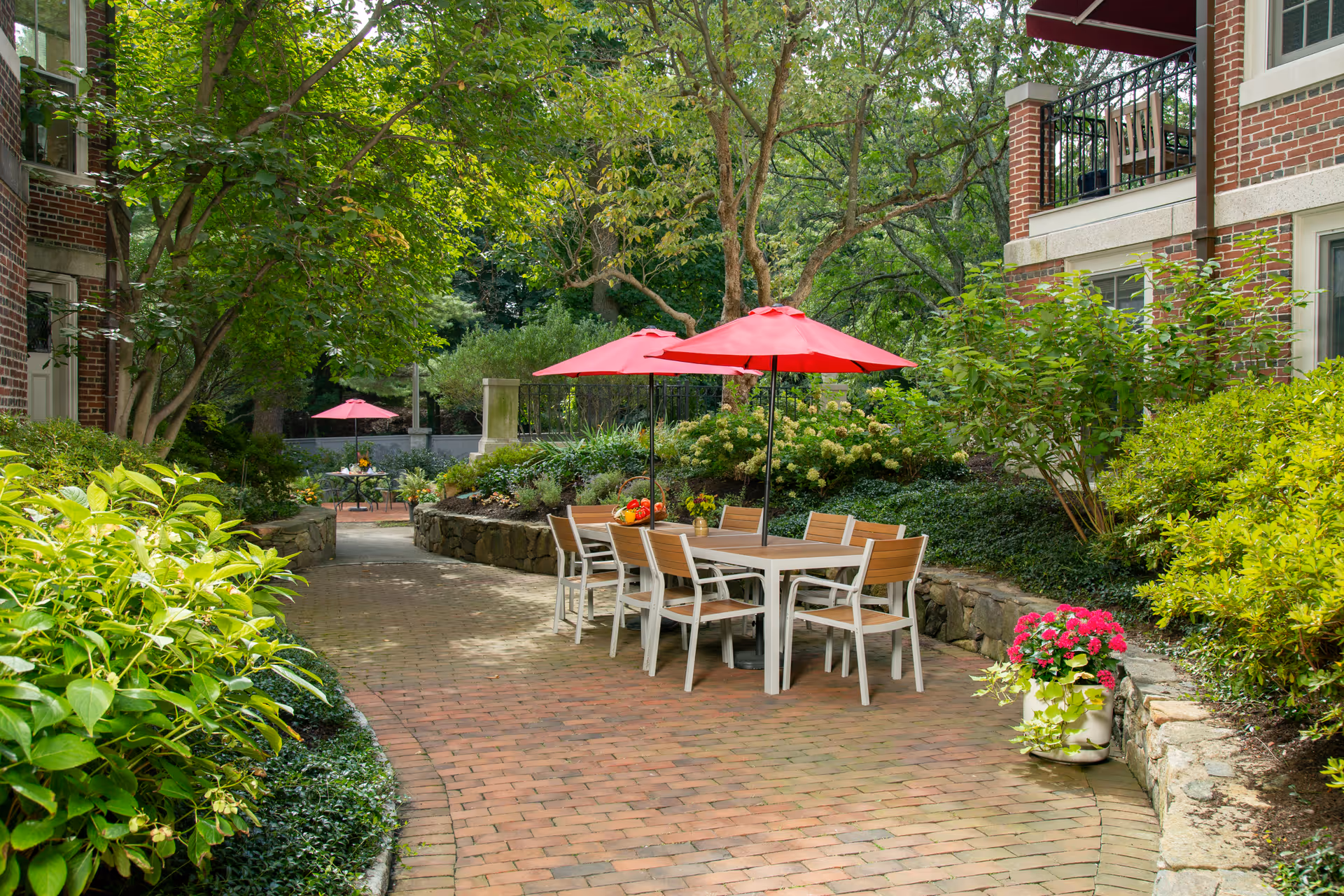 Outdoor patio area with brick pathway surrounded by lush green plants and trees. There are two tables with red umbrellas and wooden chairs, one closer to the camera and one further away. The setting is peaceful and inviting, adjacent to a brick building with balconies.