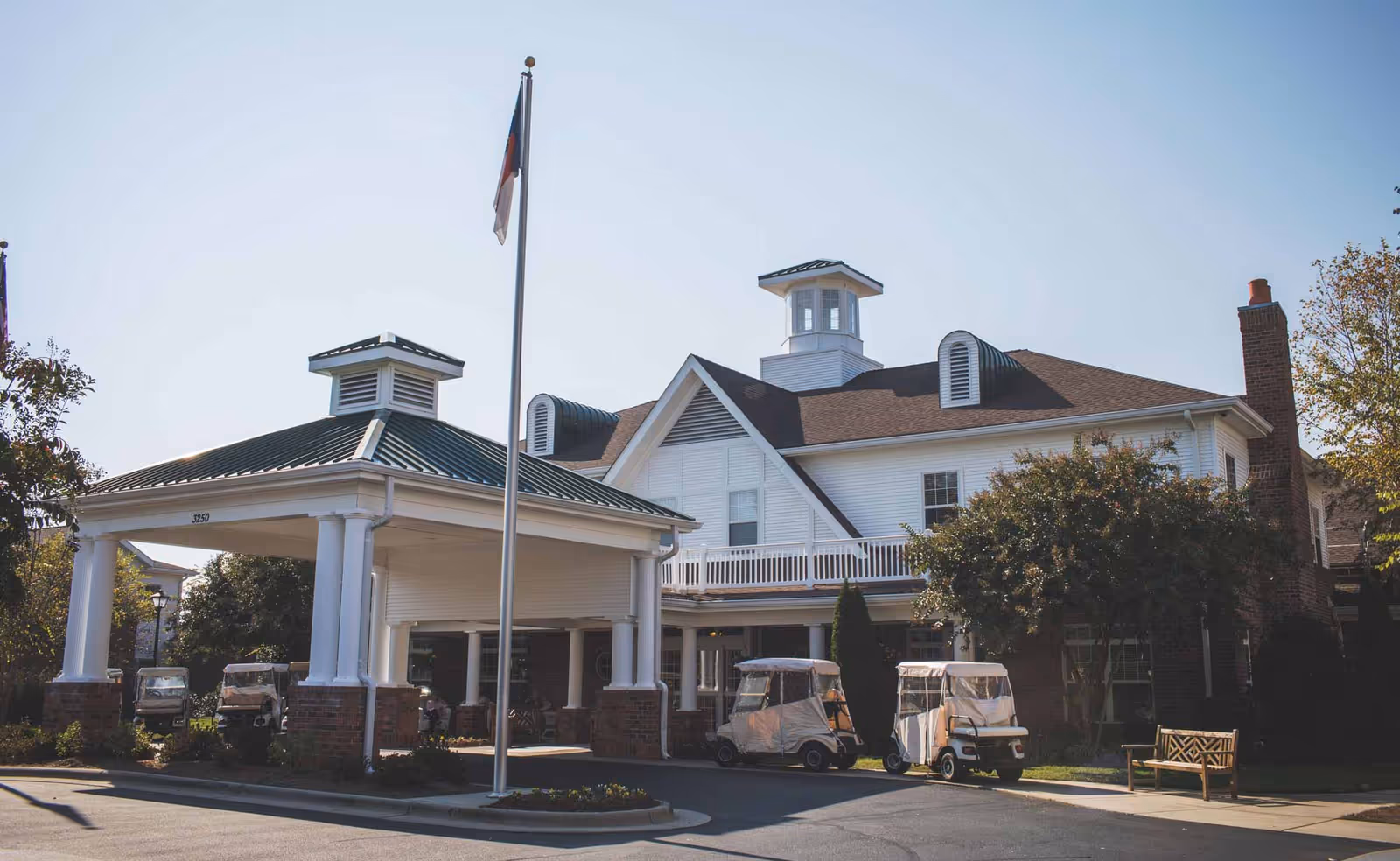 Exterior view of a senior living facility building with a covered entrance supported by white columns and brick bases. There are two golf carts parked near the entrance, a flagpole with a flag, trees, and a wooden bench on the right side. The building has white siding, multiple windows, and a distinctive cupola on the roof.