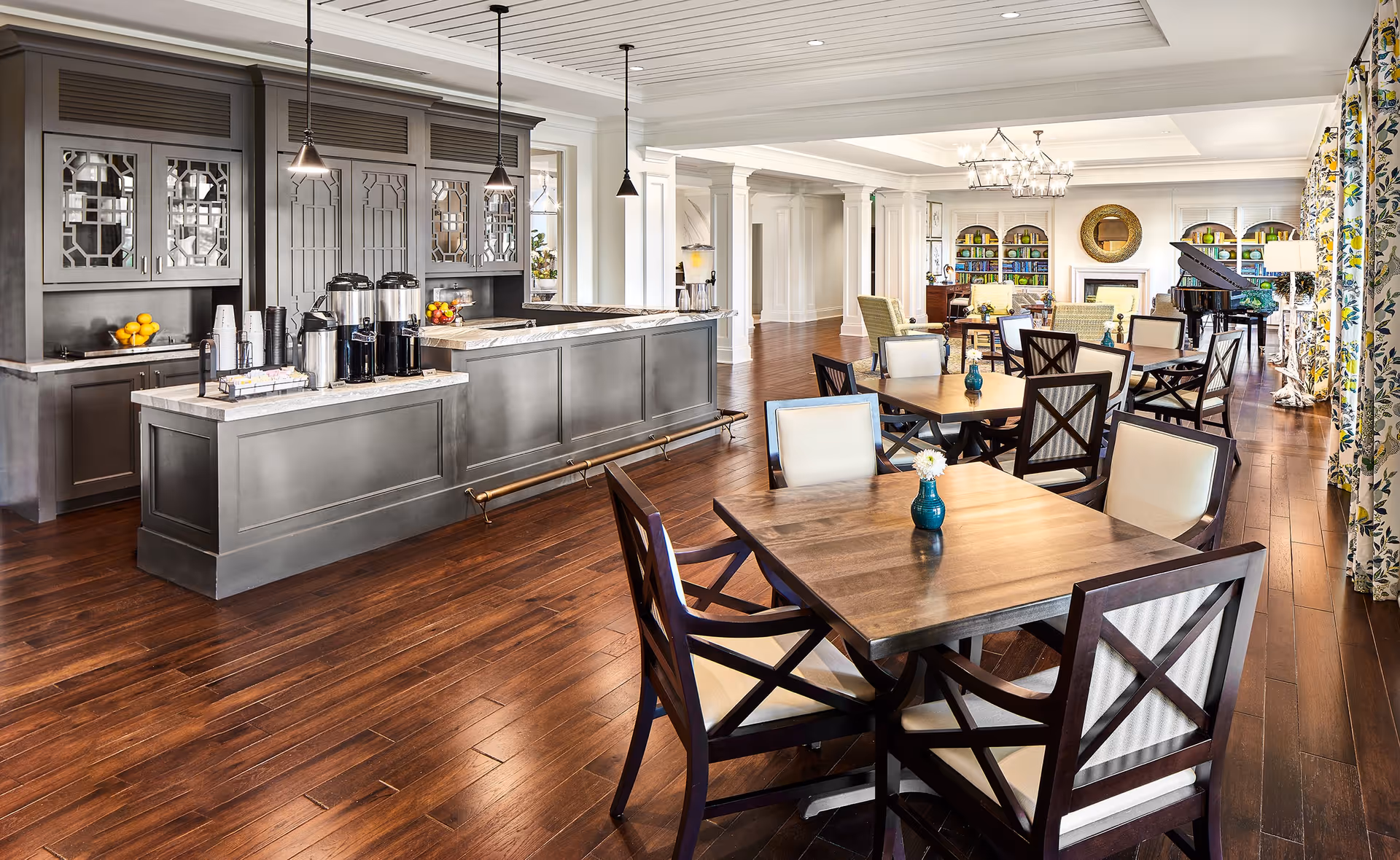 A spacious and elegant dining area with wooden floors, multiple tables and chairs, a coffee and beverage station with pendant lights, and a cozy seating area with bookshelves, a fireplace, and a grand piano in the background. The room is well-lit with natural light coming through large windows with floral curtains.