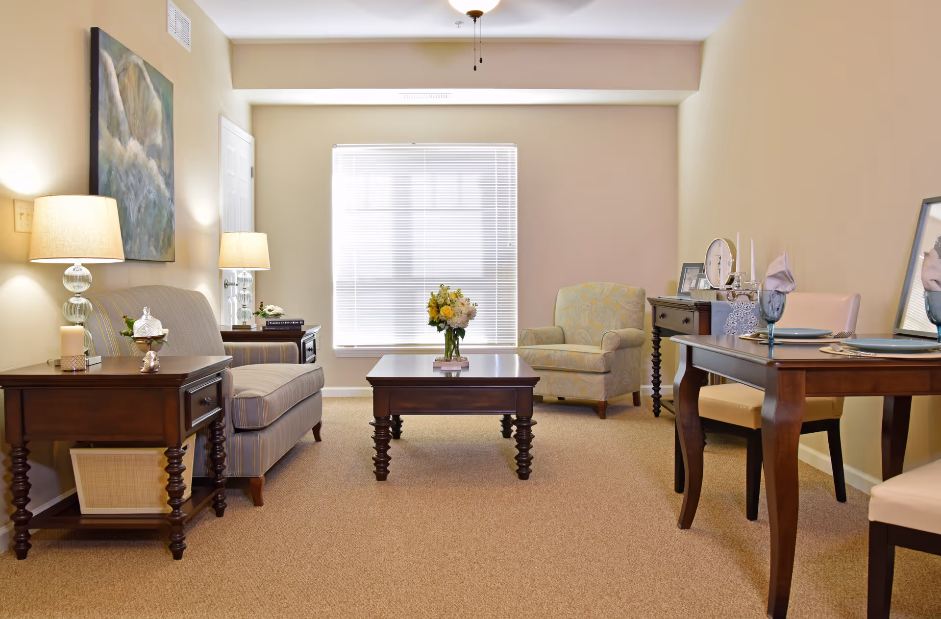 A cozy living room in Irene Woods Assisted Living featuring a striped loveseat with two lamps on side tables, a wooden coffee table with a flower vase, a patterned armchair, and a dining table set with plates, glasses, and napkins. The room has beige walls and carpet, with a large window covered by blinds letting in natural light.