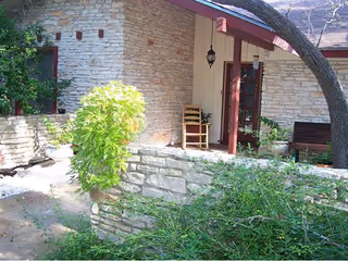 Outdoor patio area of a building with stone walls, a wooden chair, a hanging lantern, and various green plants and bushes surrounding the space.