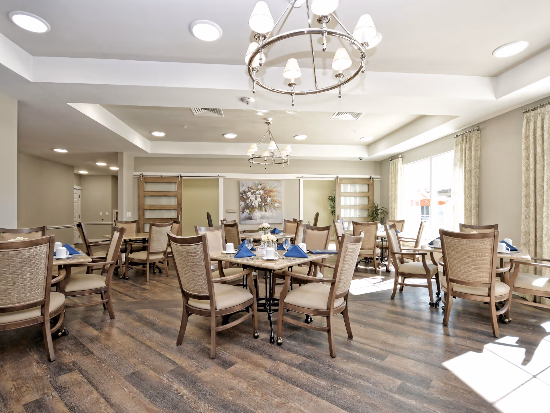 Bright and spacious dining room with multiple wooden tables and chairs arranged neatly. Each table is set with blue napkins, white cups, and glassware. The room features large windows with curtains allowing natural light to fill the space, wooden flooring, and modern ceiling lights including chandeliers.