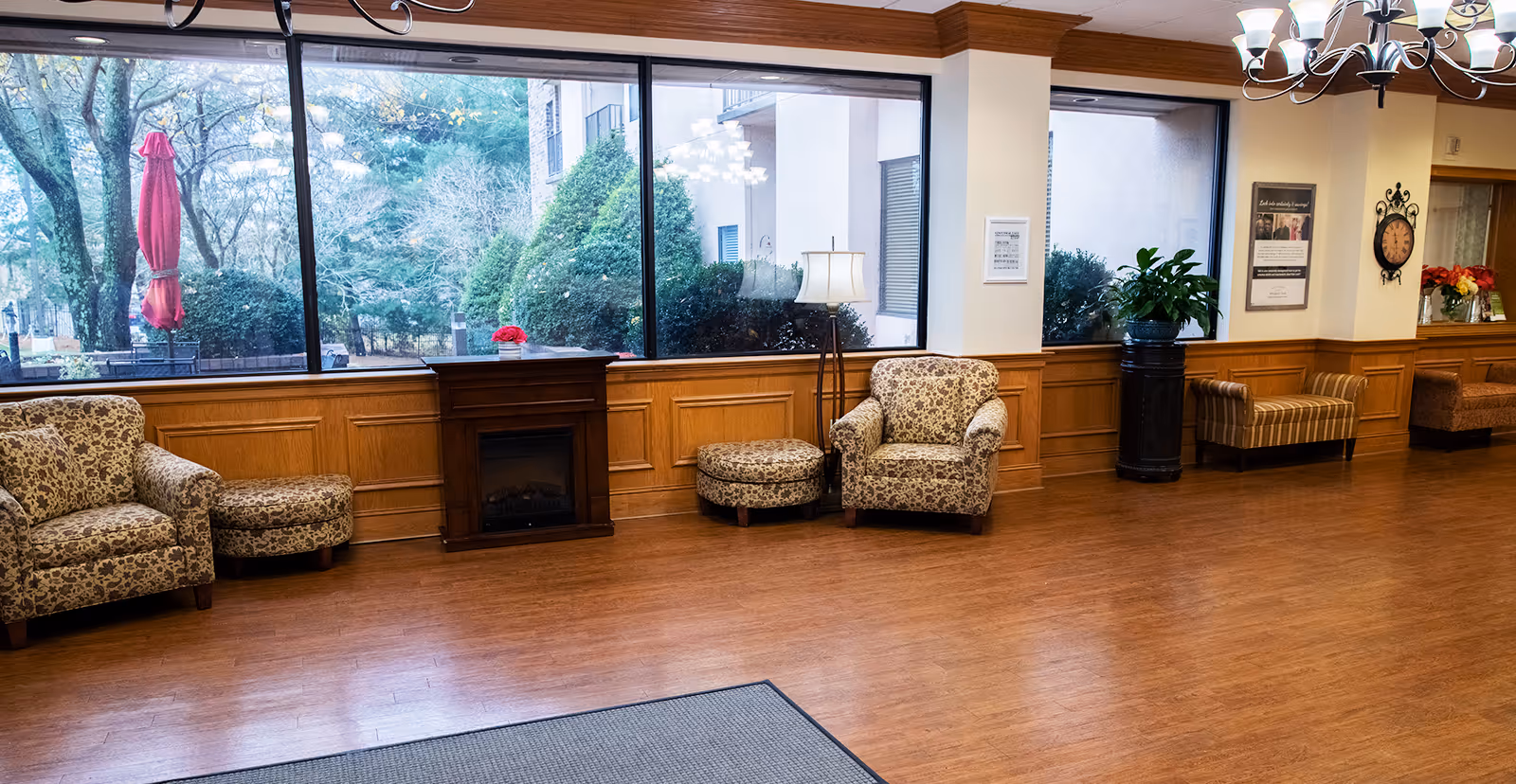 Bright communal sitting area with patterned armchairs and ottomans, wood-paneled walls, large windows looking out to trees, and a chandelier.