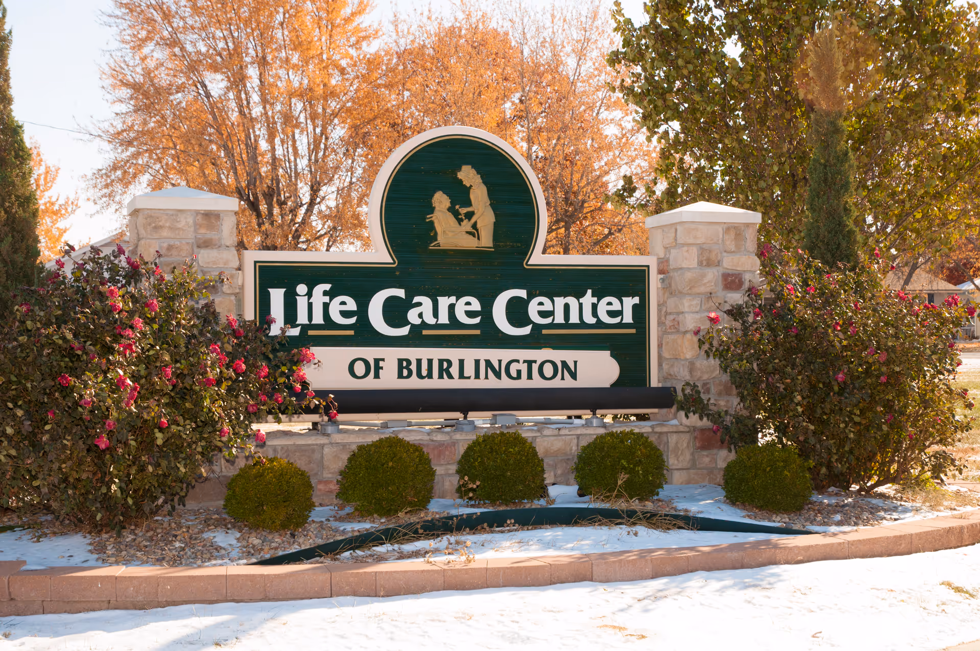 Outdoor sign for Life Care Center of Burlington surrounded by bushes with red flowers and small green shrubs, with autumn trees and a light layer of snow on the ground.