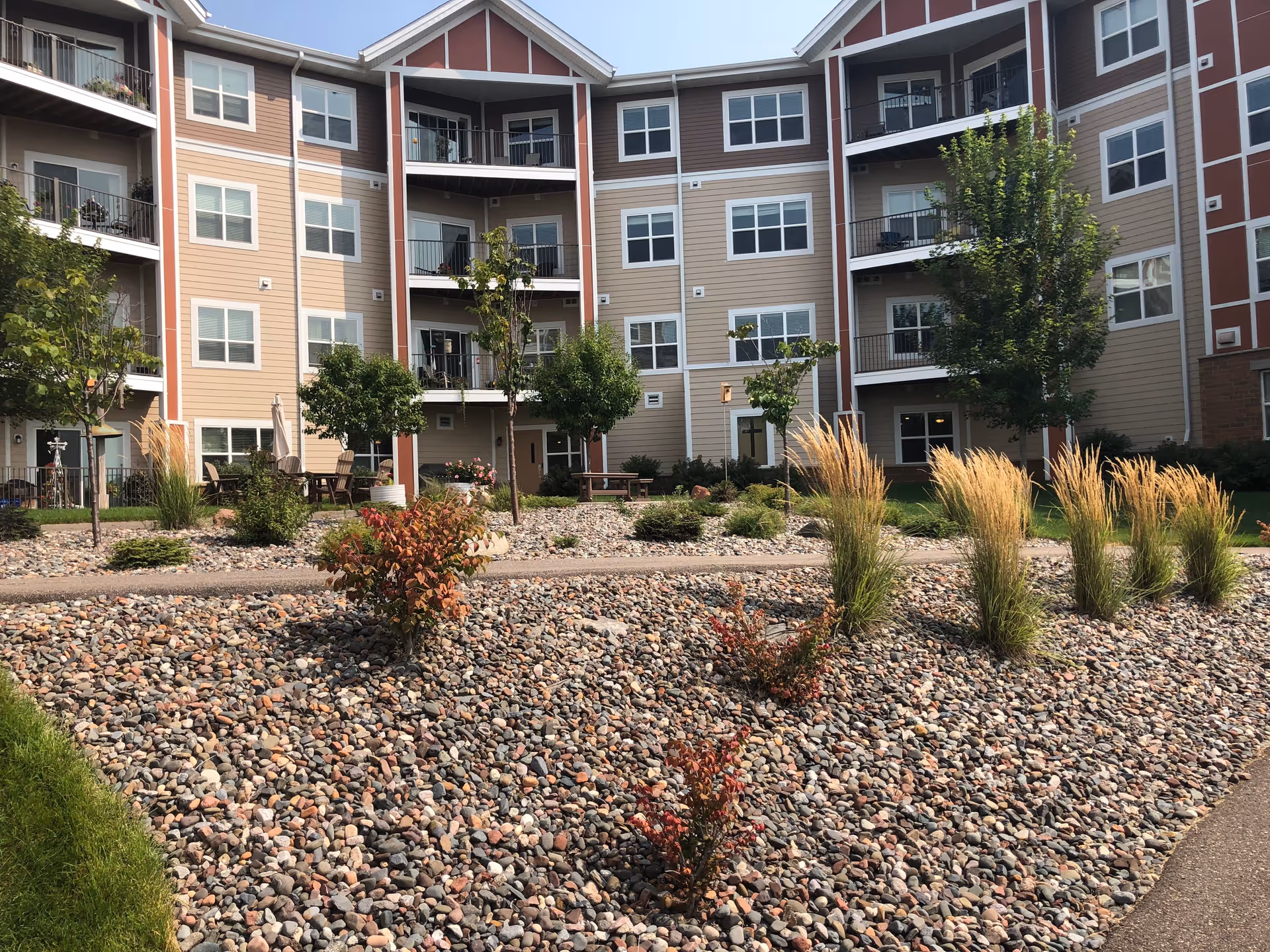 Outdoor landscaped courtyard area of a senior living facility with a variety of small trees, shrubs, and ornamental grasses planted in a bed of decorative rocks. The courtyard is surrounded by a multi-story beige and brown building with balconies and windows. There are outdoor chairs and tables visible near the building.