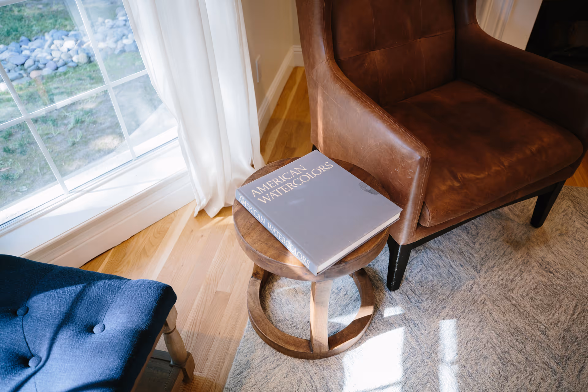 A cozy corner with a brown leather armchair, a small round wooden side table holding a book titled 'American Watercolors', a blue cushioned chair, and a window with white curtains letting in natural light.