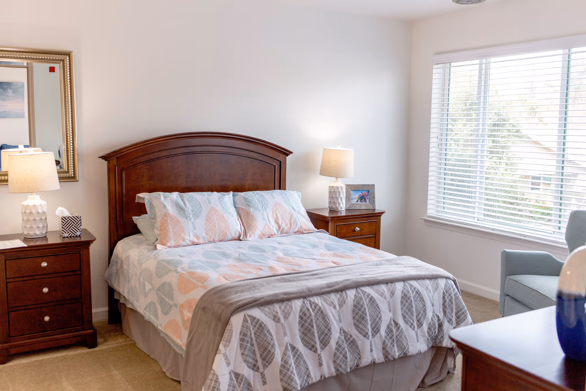 A bright and cozy bedroom with a wooden bed frame, a bed made with patterned sheets and pillows in pastel colors. There are two wooden nightstands on either side of the bed, each with a lamp. A large window with white blinds lets in natural light, and a light blue armchair is positioned near the window. A framed picture and a mirror hang on the walls.