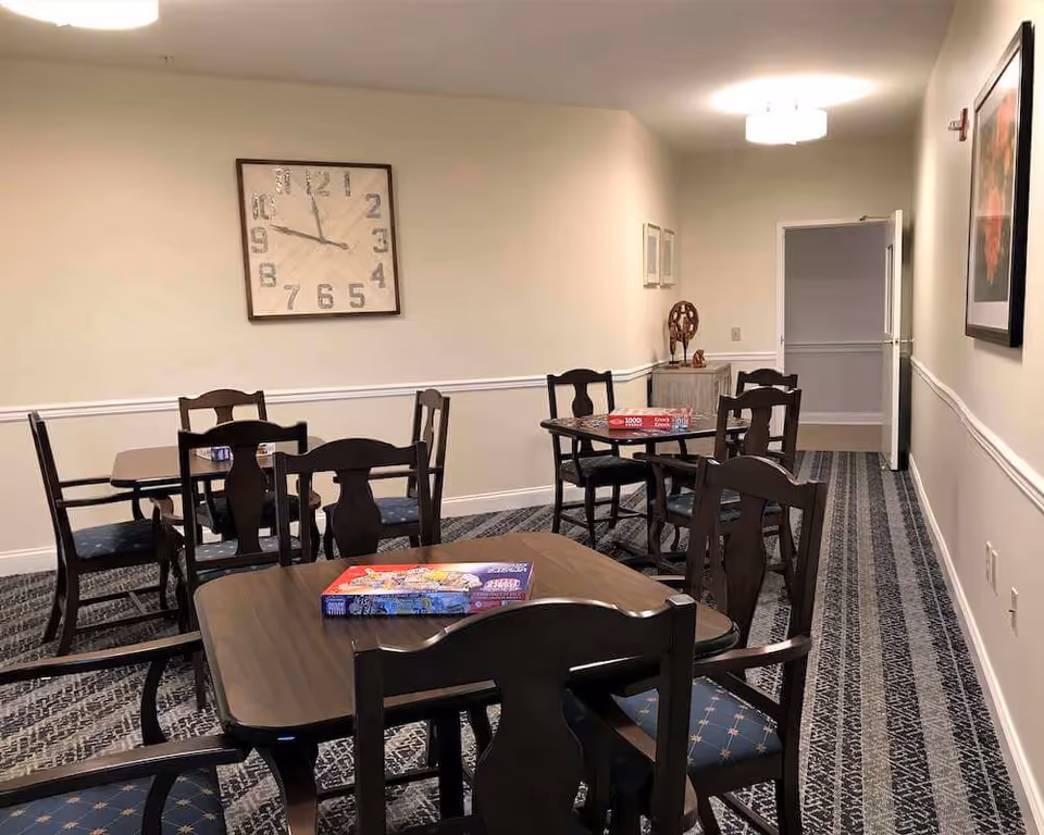 Communal activity room with several wooden tables and chairs, a large wall clock, and board games on the tables.