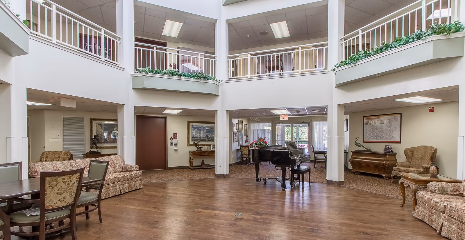 Spacious senior living facility common area with a grand piano in the center, surrounded by comfortable seating including floral patterned sofas and armchairs. The room has a high ceiling with a second-floor balcony adorned with green plants. Large windows and glass doors provide natural light and a view of the outdoor area.