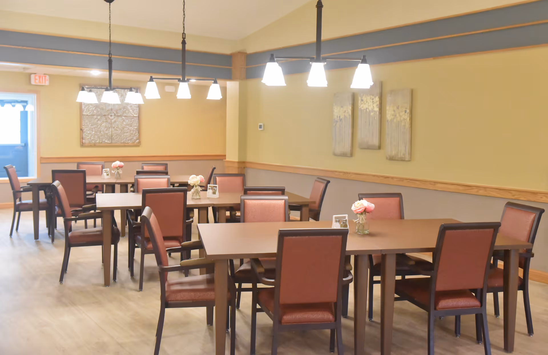 Dining room with multiple square tables and red-upholstered chairs arranged under hanging lights.