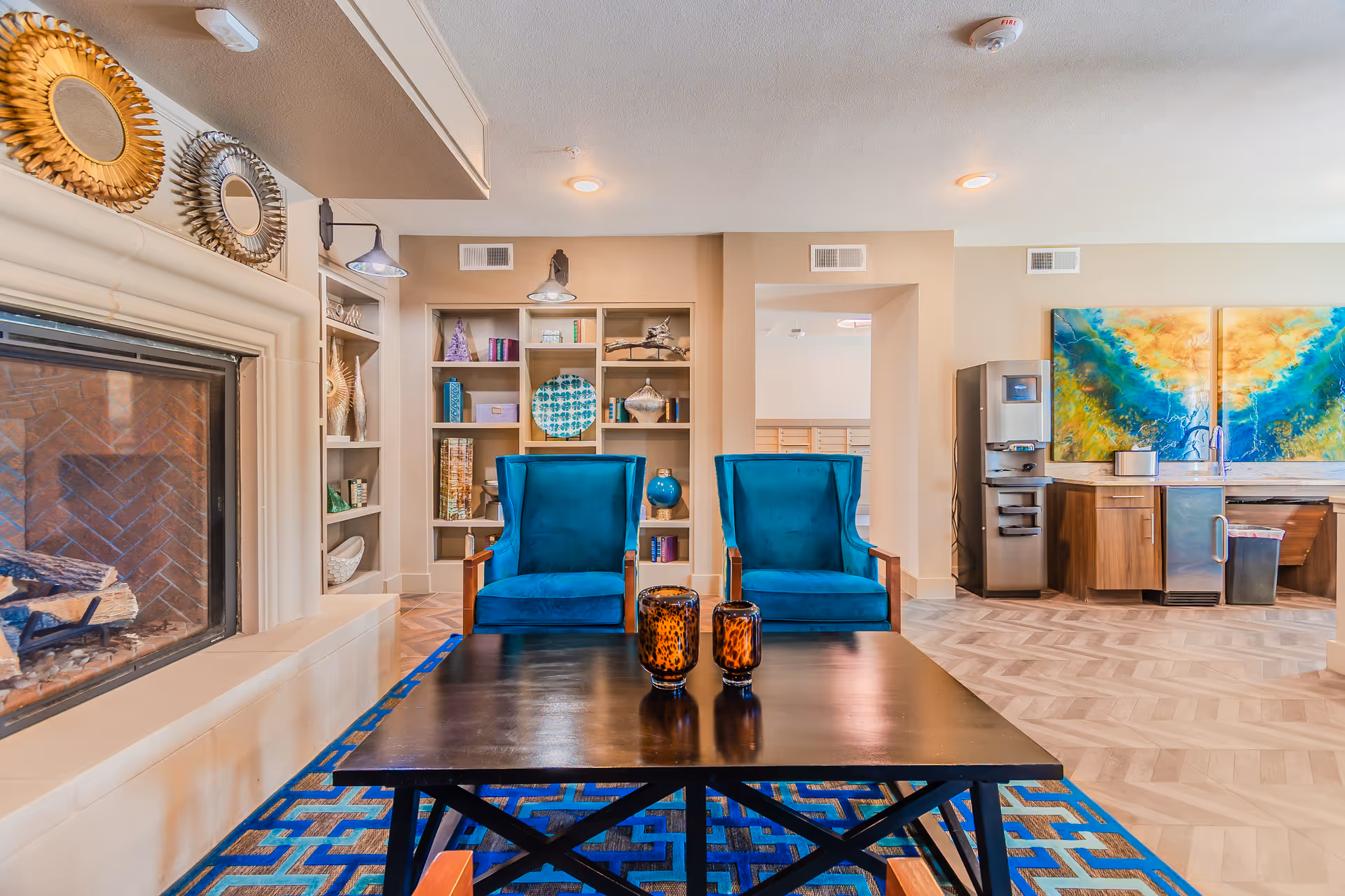 A cozy living room area featuring two blue velvet armchairs facing a black coffee table with two decorative amber glass candle holders. To the left is a fireplace with a white mantel adorned with two round sunburst mirrors. Behind the chairs is a built-in shelving unit with various decorative items and books. To the right, there is a kitchenette area with a water dispenser, sink, and colorful abstract artwork on the wall.