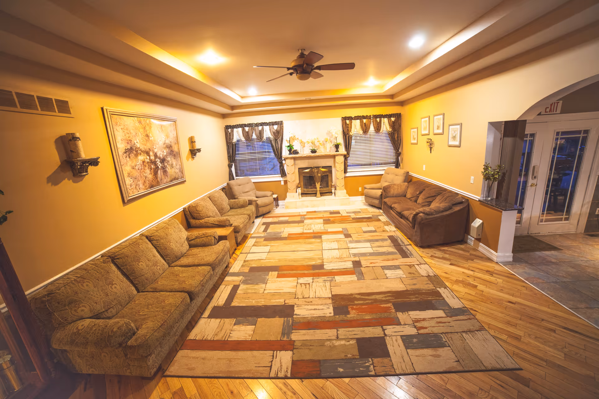 A cozy living room with multiple upholstered sofas and armchairs arranged around a large patterned area rug. The room features a fireplace centered between two windows with curtains, warm yellow walls, wooden flooring, a ceiling fan, and framed artwork on the walls. There is an exit door visible on the right side.