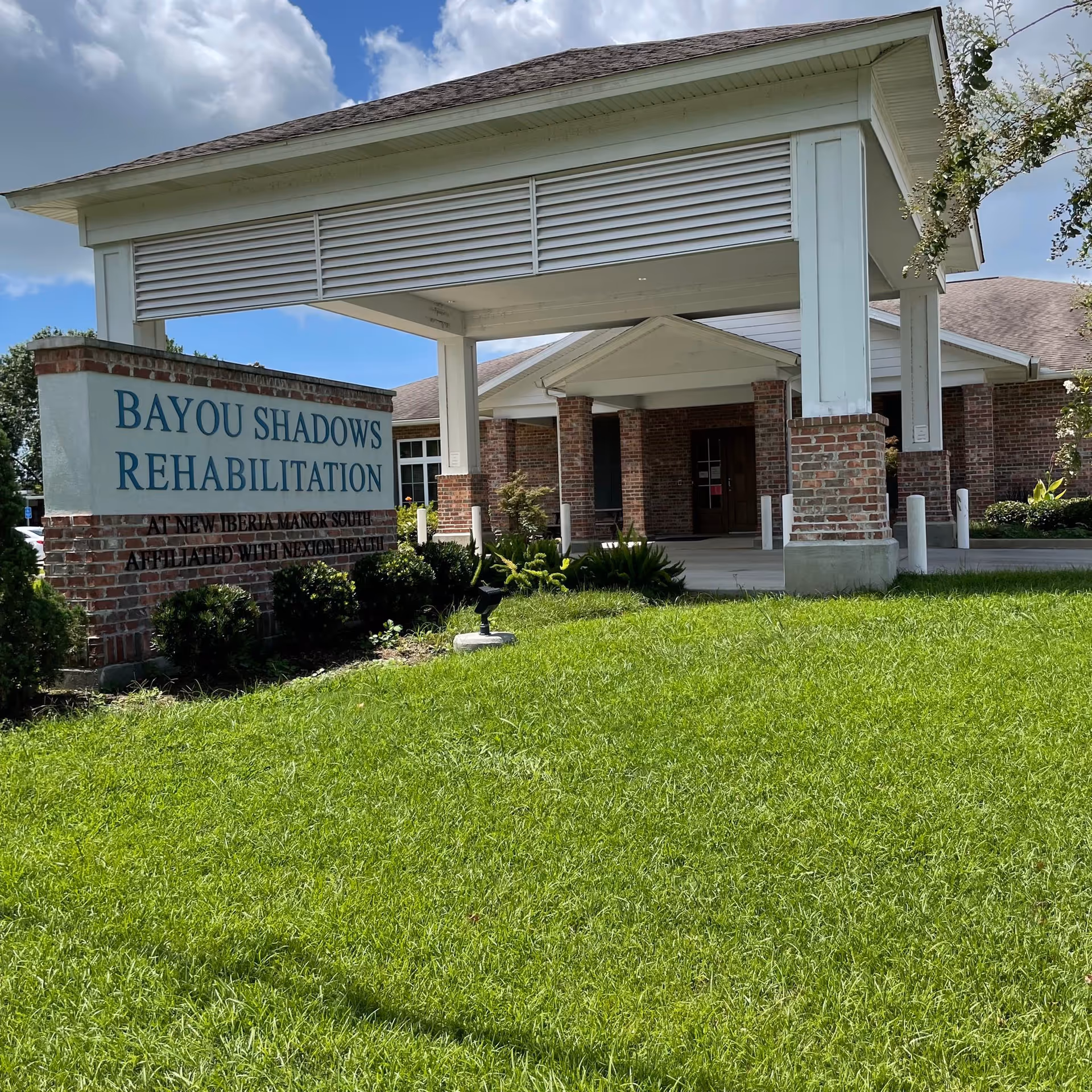 Front entrance canopy and sign reading 'Bayou Shadows Rehabilitation' at New Iberia Manor South with a grassy lawn in the foreground.