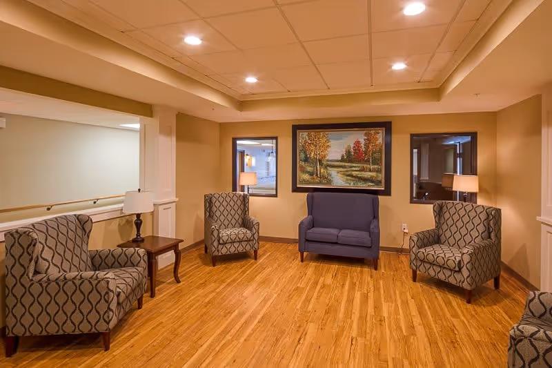A cozy sitting area in a senior living facility with four patterned armchairs and one blue loveseat arranged around a wooden floor. There are two small side tables with lamps, beige walls, and a large framed painting of an autumn landscape on the wall.