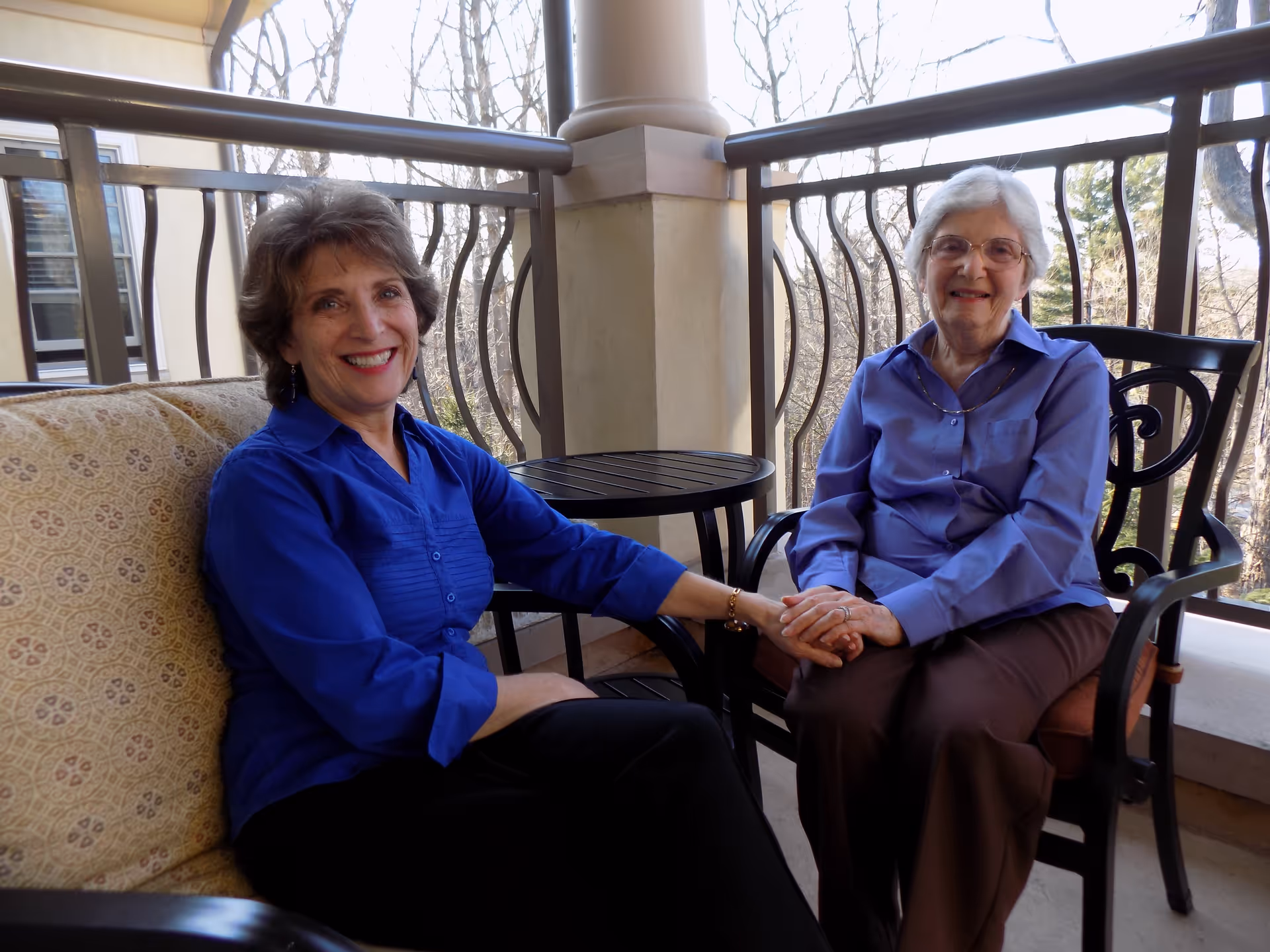 Two women sitting on a balcony holding hands and smiling. One woman is seated on a cushioned bench, wearing a blue shirt and black pants, while the other woman is seated on a chair, wearing a light blue shirt and brown pants. The balcony has a metal railing and a small round table between them, with trees visible in the background.