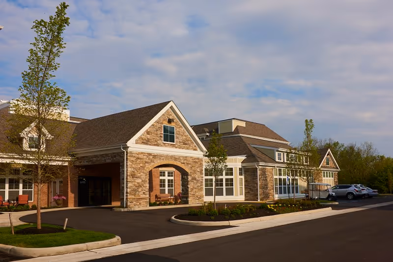 Exterior view of a single-story building with stone and brick facade, large windows, and a covered entrance. There are small trees and landscaped areas with grass and flowers in front of the building, along with a paved driveway and parked cars. The sky is partly cloudy.