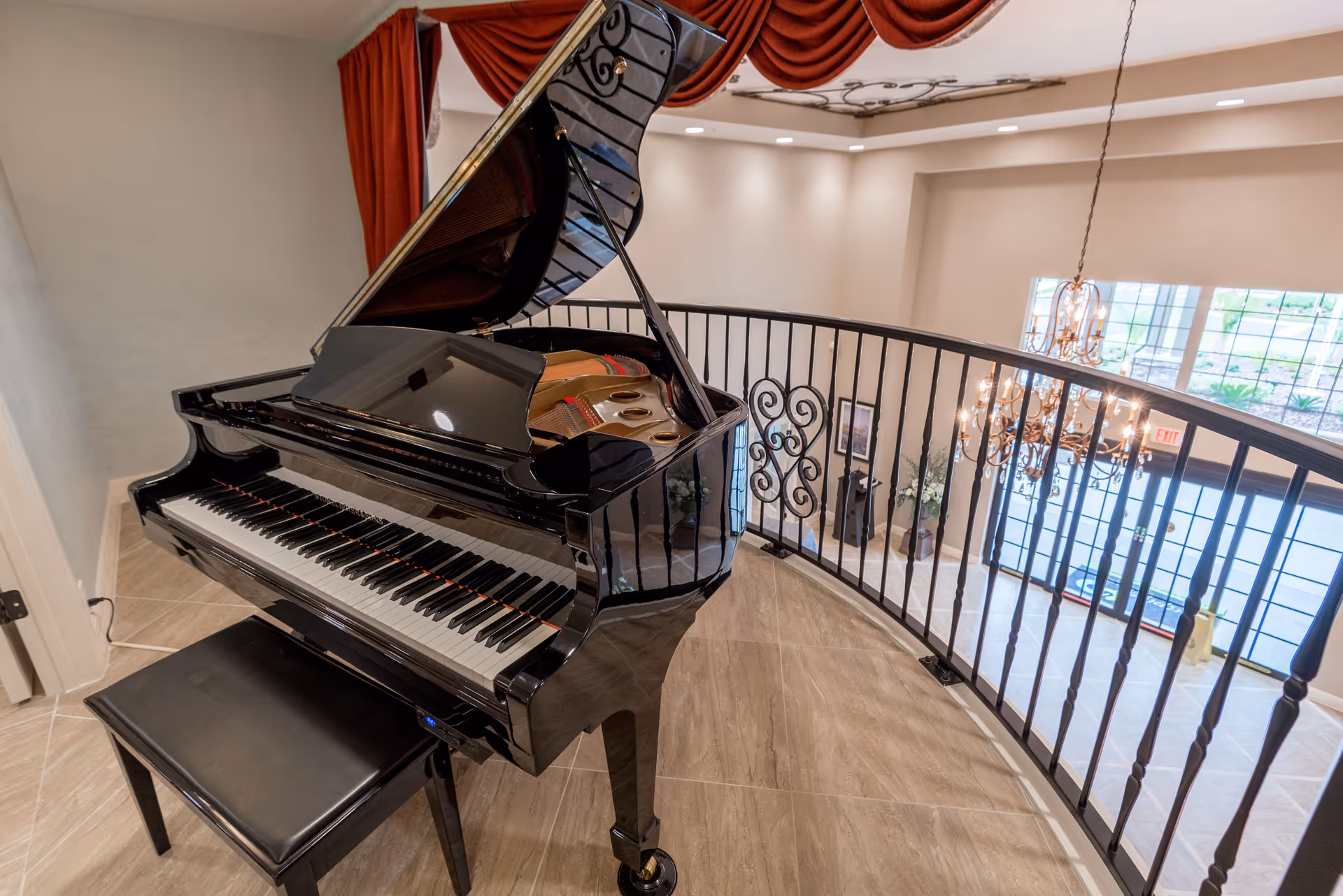 A black grand piano with its lid open is placed on a tiled floor near a curved black metal railing overlooking a spacious, well-lit lobby area with large windows and a chandelier. Red drapes hang above the piano.
