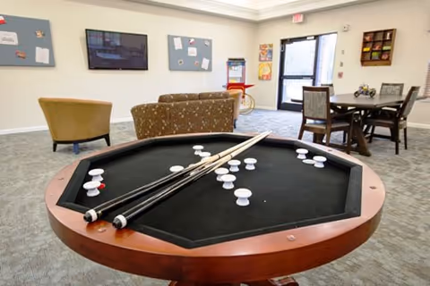 A recreational room with a wooden octagonal pool table in the foreground, featuring several white and red pool balls and three pool cues resting on it. In the background, there are upholstered chairs and a sofa facing a wall-mounted TV, bulletin boards on the walls, a table with chairs, and a door leading outside.