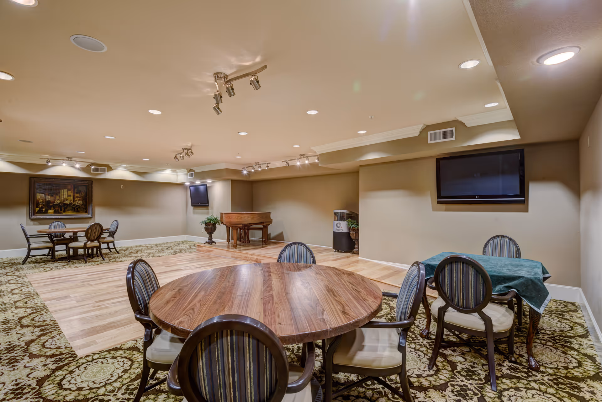 A spacious common room with multiple round wooden tables surrounded by striped cushioned chairs. The room features a patterned carpet with a wooden floor section in the center, a grand piano on a small raised platform, two wall-mounted flat screen TVs, and a large framed painting on one wall. The ceiling has recessed lighting and track lights.