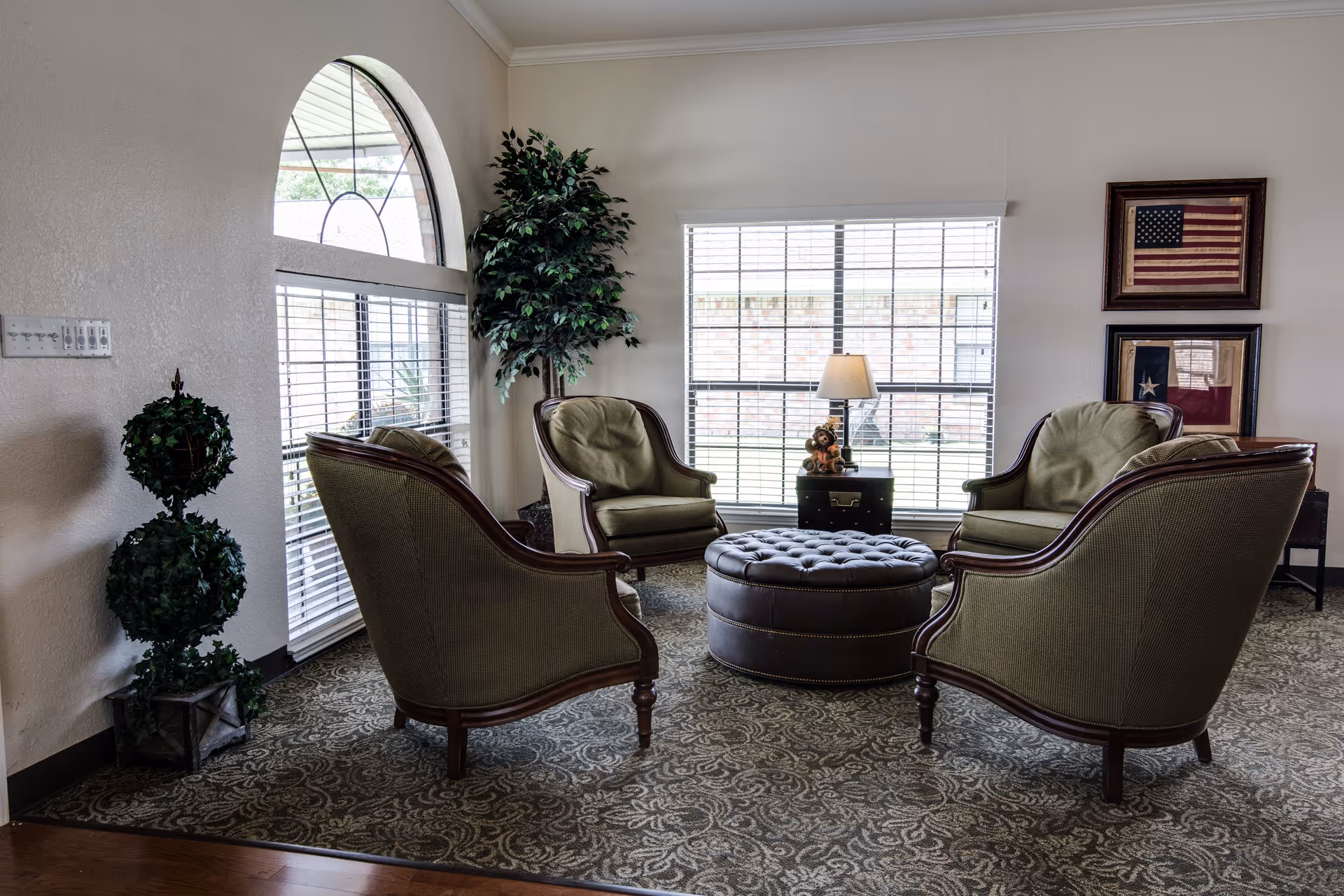 A cozy sitting area with four green upholstered armchairs arranged around a round, tufted ottoman. The space features a large window with blinds, a small side table with a lamp and decorative items, two framed flags on the wall, and two artificial plants. The floor is covered with a patterned carpet.