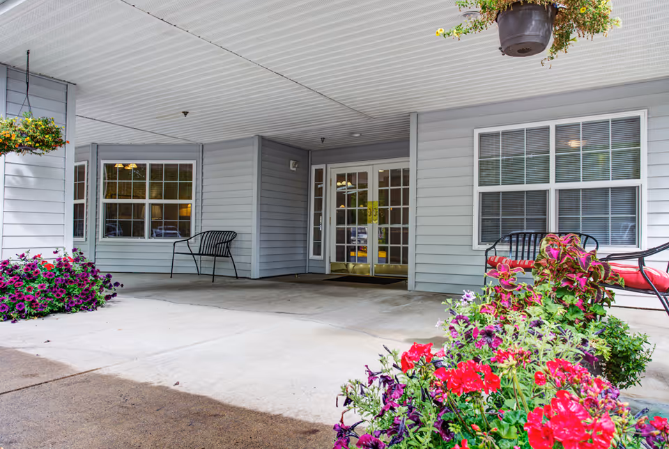 Covered entrance area of Creekside Place Memory Care facility with double glass doors, gray siding, two black metal benches, and vibrant flower arrangements in the foreground and hanging planters.