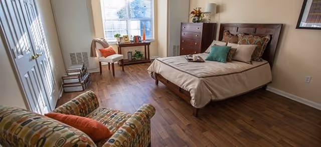 Well-lit bedroom with a bed, patterned armchair, wooden dresser, and a window with blinds.