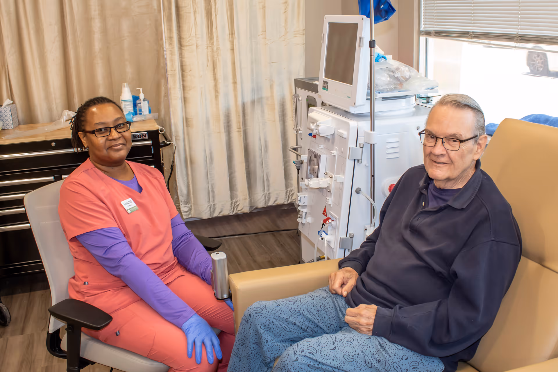 A healthcare worker in pink scrubs and blue gloves sits next to an elderly man receiving dialysis treatment in a medical facility. The man is seated in a beige recliner chair connected to a dialysis machine. Both individuals are smiling and looking at the camera.