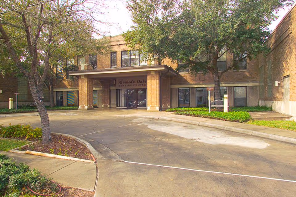 Front exterior view of Alameda Oaks Nursing Center showing a two-story brick building with a covered entrance, surrounded by trees and landscaped greenery.