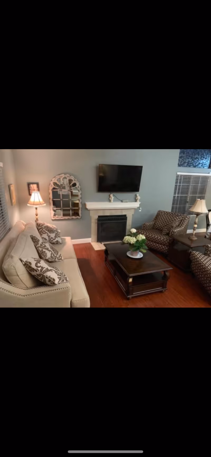 A cozy living room with a beige sofa adorned with patterned cushions, two patterned armchairs, a dark wooden coffee table with a flower arrangement, a fireplace with decorative items on the mantel, a wall-mounted flat-screen TV, two table lamps, and a decorative mirror on the wall.