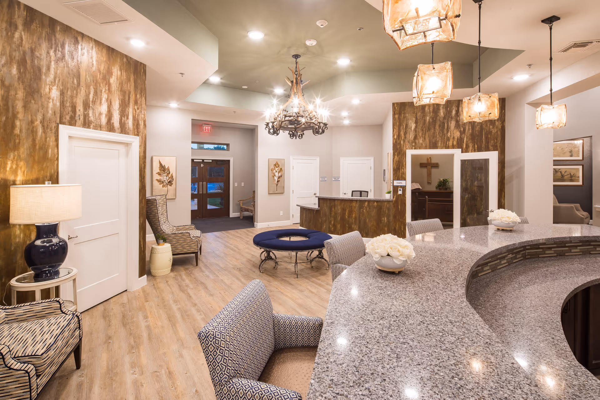 A spacious and warmly lit interior of a senior living facility reception area with a curved granite countertop, several upholstered chairs, a chandelier, pendant lights, decorative wall art, and a wooden cross visible in a room behind glass doors.