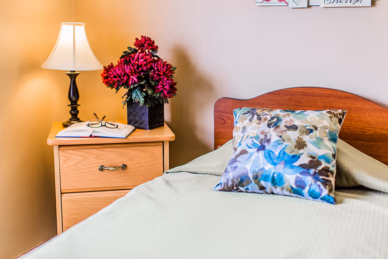 A bedside table with a lamp, open book and glasses, and a vase of red flowers next to a neatly made bed with a patterned pillow.