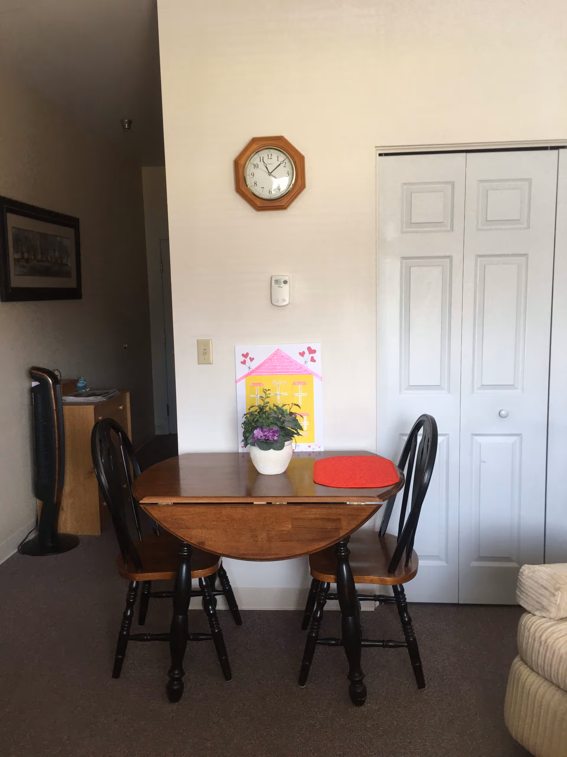 A small wooden dining table with two chairs in a room. On the table is a potted plant and a red placemat. Behind the table is a white wall with a clock, a thermostat, and a colorful drawing of a house. To the right is a closed white closet door, and to the left is a partial view of a desk and a standing fan.